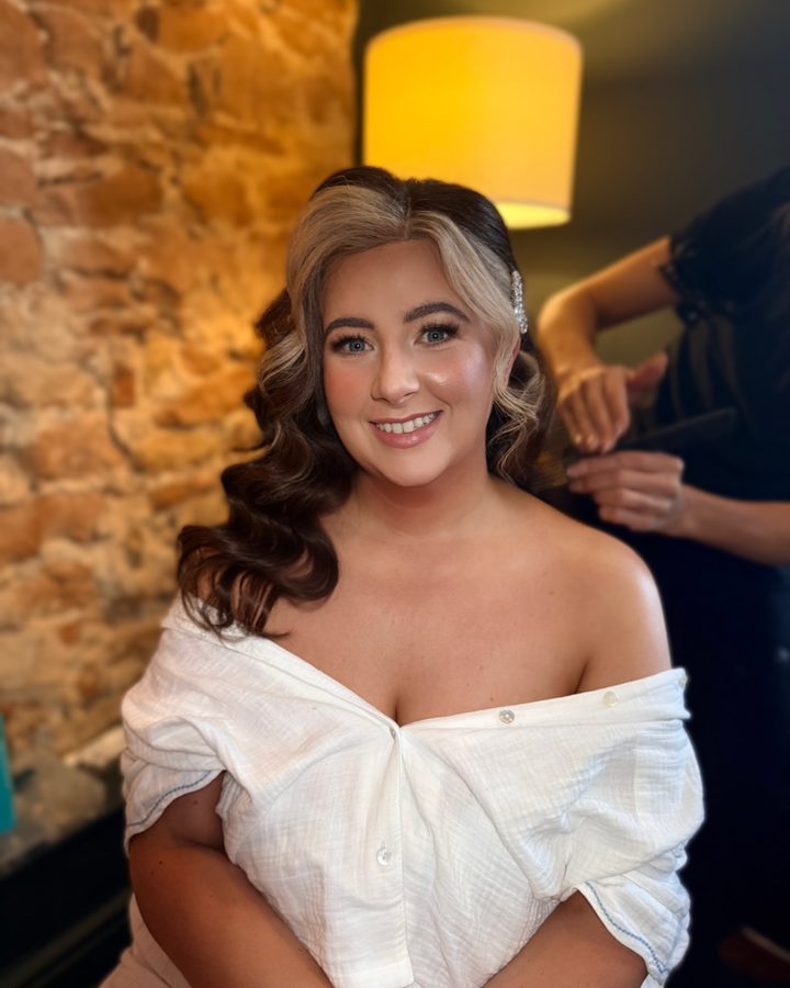 A bride with glowing bridal makeup and her hair being styled, by By.EmWebster